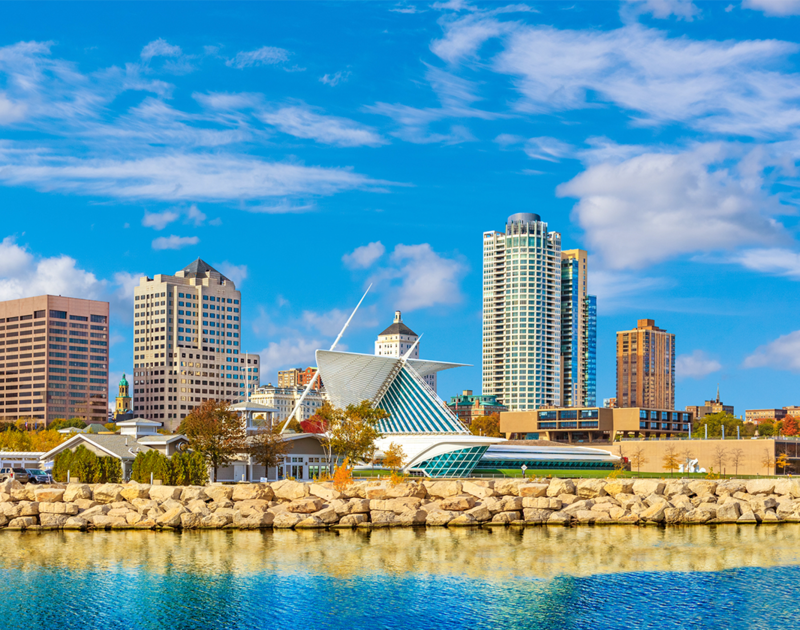 Milwaukee downtown skyline set against blue sky is reflected in the waters of Lake Michigan in foreground.